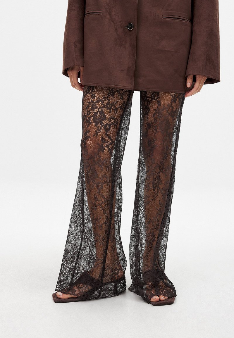 Pantaloni a zampa d'elefante in pizzo nero con motivo floreale, tessuto trasparente e aperture delle gambe ampie, abbinati a sandali scivolati marroni.