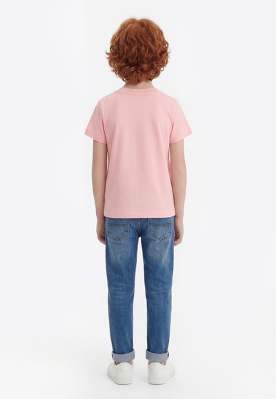 Παιδί με σγουρά κόκκινα μαλλιά, φορώντας ροζ μπλουζάκι με κοντά μανίκια, μπλε τζιν παντελόνι με γυρισμένα μπατζάκια και λευκά αθλητικά παπούτσια, στέκεται με την πλάτη στραμμένη σε λευκό φόντο.