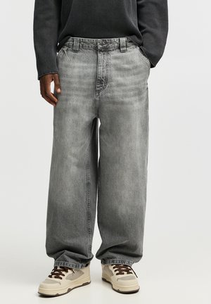 Personne portant un jean gris ample, un haut à manches longues foncé et des baskets beige avec des lacets marron, debout devant un fond uni.