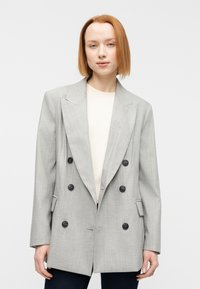 Blazer gris croisé avec de grands boutons noirs, des revers à crans, et un tissu texturé. Porté sur un haut crème et un pantalon foncé.