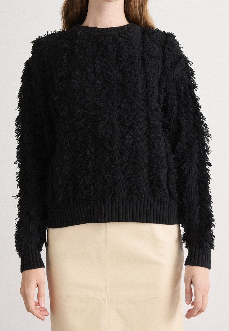 Femme portant un pull en maille texturée noir avec des rayures verticales en relief et une jupe beige, debout devant un fond uni.