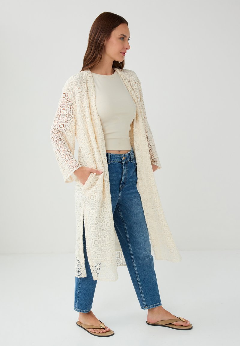 Der Spitzenkimono in Creme verfügt über ein geometrisches Muster, einen offenen Schnitt, lange Ärmel, Taschen und seitliche Schlitze und wird mit Jeans und Sandalen kombiniert.