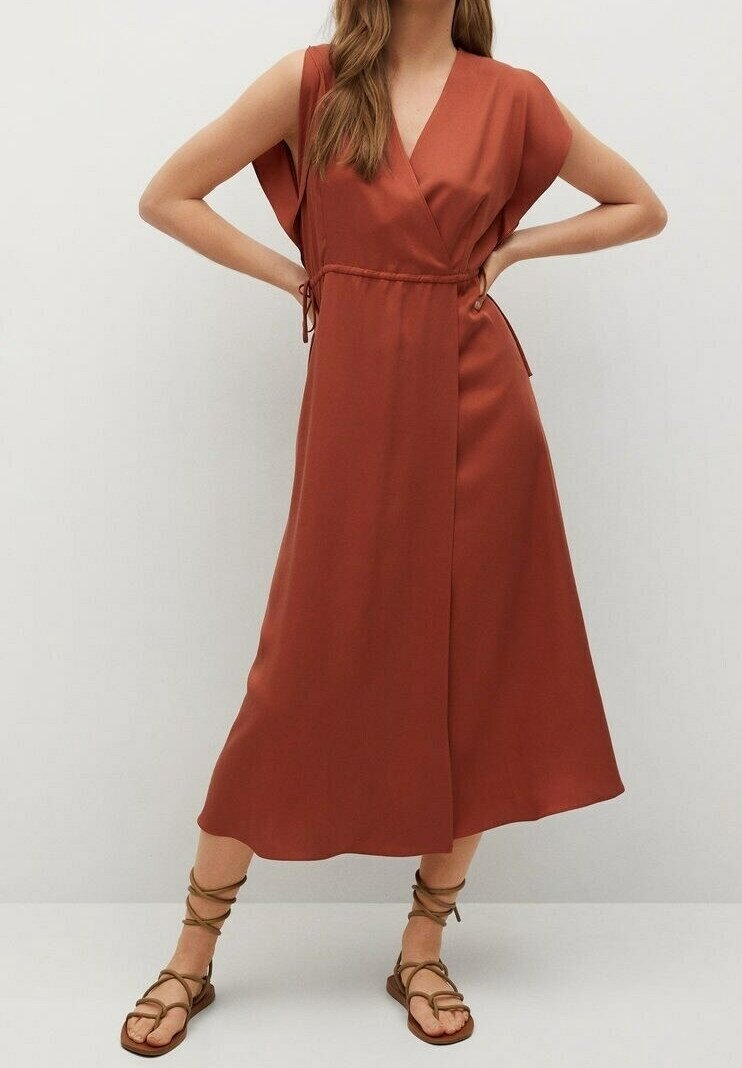 Femme portant une robe midi portefeuille sans manches couleur rouille avec taille froncée et sandales plates marron à lacets, debout les mains sur les hanches.
