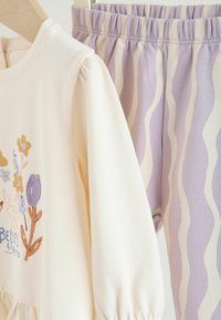 Krém színű felső virág hímzéssel, amelyet levendula színű leggings követ, hullámos csíkkal. Mindkét ruhadarab puha, rugalmas anyagból készült, redőzött részletekkel.