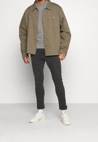 Chaqueta camisa verde oliva con dos bolsillos en el pecho, camiseta gris debajo, jeans negros ajustados y zapatillas blancas. Tejido plano y texturizado.