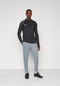 Black long-sleeve athletic shirt with a quarter-zip and grey accents, paired with light grey pants featuring a red logo, and black athletic shoes.