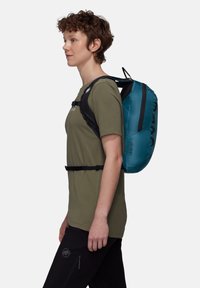 Νεαρό άτομο με πουκάμισο χακί και μαύρο παντελόνι, που φοράει μια τυρκουάζ τσάντα πλάτης με μαύρα λουριά, στέκεται προφίλ σε λευκό φόντο.