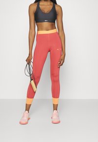 Reggiseno sportivo nero, leggings rossi con accenti arancioni e sneaker rosa. I leggings presentano un logo Puma; si sta tenendo una corda per saltare. Texture liscia.