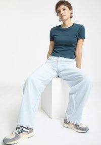 Teal eng geschnittenes T-Shirt, hellblaue weit geschnittene Jeans und graue Sneakers mit Akzenten. Model sitzt auf einem Würfel vor einem schlichten Hintergrund.