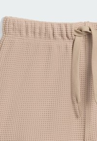 Primer plano de la cintura de unos pantalones de punto waffle beige con elástico y cordón ajustado sobre un fondo claro.