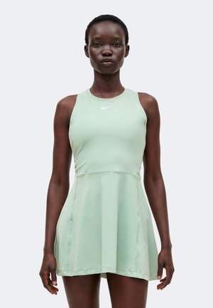 Frau steht frontal, trägt ein ärmelloses hellgrünes Nike-Sportkleid mit ausgestelltem Rock vor einfarbigem Hintergrund.