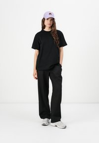 T-shirt en coton noir associé à un pantalon noir ample. Une casquette lavande avec un logo floral complète la tenue, accompagnée de baskets grises.