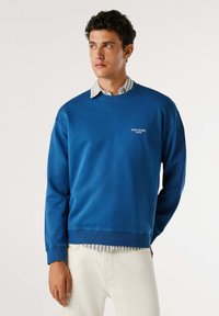 Felpa blu a girocollo realizzata in materiale morbido, con polsini e orlo a coste, e un logo bianco sul petto. Indossata con una camicia a righe sotto.