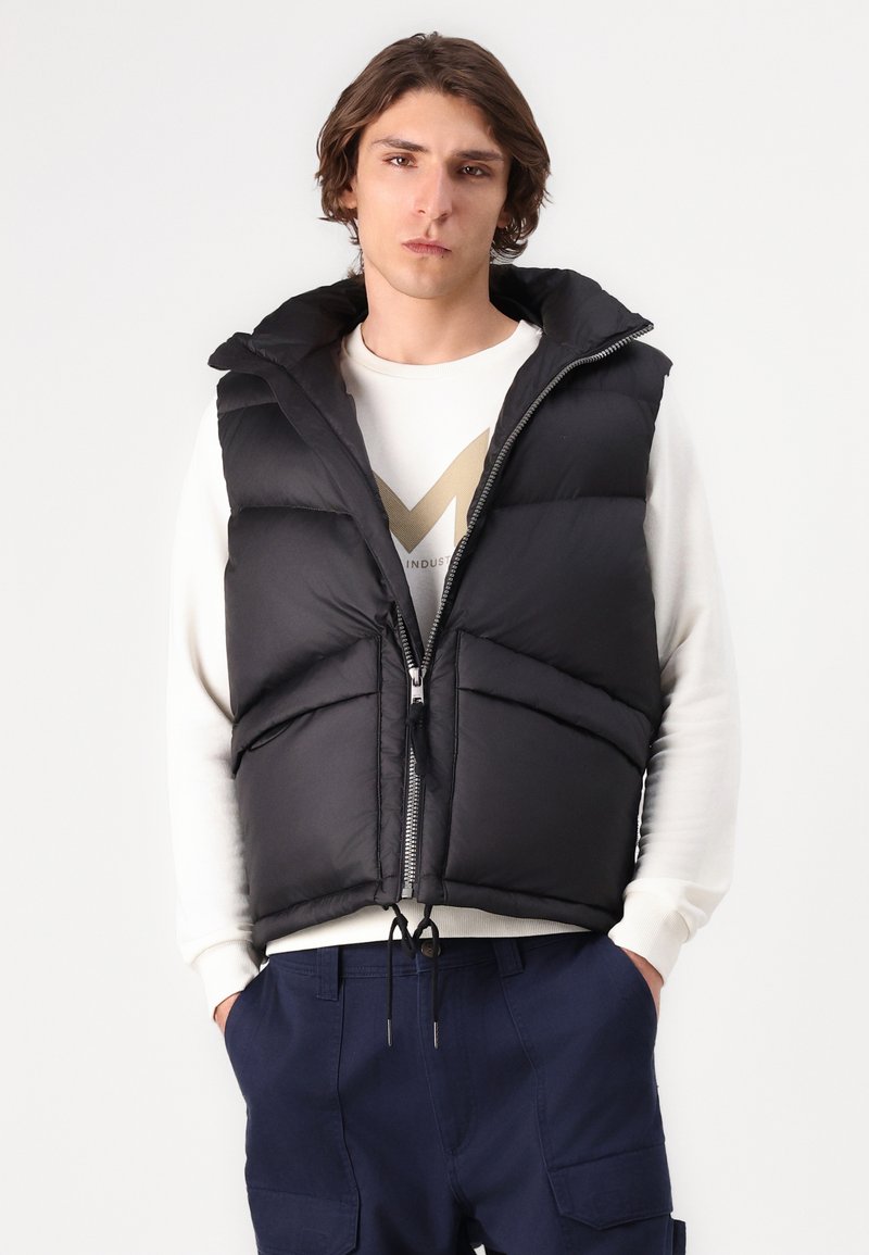 Gilet rembourré noir avec un col haut, fermeture éclair à l'avant et deux grandes poches avant, porté sur un sweat-shirt blanc et un pantalon bleu marine.