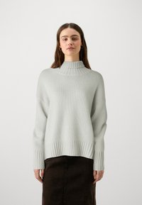 Pull en maille gris clair avec col roulé, doté d'un ourlet et de poignets côtelés, offrant une coupe décontractée et une texture douce, associé à une jupe marron foncé.