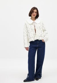 Veste blanche en quilté avec broderie florale, grand col blanc et fermeture à l'avant par lien ; associée à un pantalon en denim bleu large avec lanières aux chevilles.