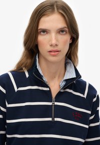 Jeune femme aux cheveux châtains clairs portant un pull zippé à rayures bleu marine et blanc sur une chemise à col rayée bleue et blanche.