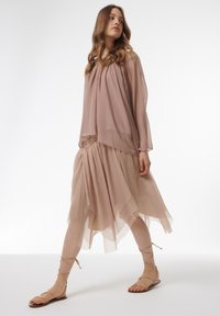 Helles rosafarbenes, transparentes Blusenshirt mit plissiertem Design, kombiniert mit einem fließenden, geschichteten nudefarbenen Rock. Strappy beige Sandalen vervollständigen das Outfit.