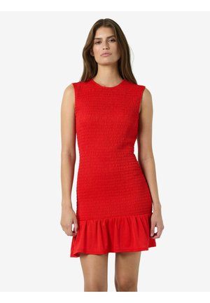 Mujer que lleva un vestido rojo sin mangas, ajustado, con tela texturizada y un dobladillo con volantes, de pie frente a un fondo claro y liso.