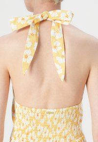 Geel bloemdress met een open rug, gekenmerkt door een halterknoopsluiting en een gerimpelde taille. De stof heeft een zachte, gestructureerde uitstraling.