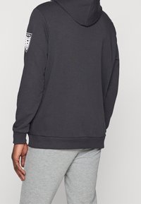 Felpa con cappuccio grigio scuro e orlo a coste, con una toppa bianca sulla spalla sinistra. Indossata con pantaloni della tuta grigi chiari.