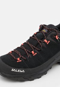 Sort vandresko med røde detaljer og struktureret mesh-øvre. Funktioner omfatter en tyk sål, mønstret snøring og fremtrædende branding.