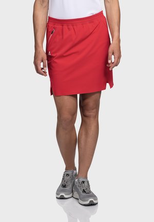Roter Sportrock mit elastischem Bund, seitlichem Reißverschluss und seitlichen Schlitzen. Getragen mit grauen Schuhen mit strukturiertem Muster.