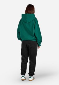 Sweatshirt verde com capuz, calças de treino pretas e ténis brancos com riscas verdes. O sweatshirt tem um corte descontraído e bolso tipo canguru.