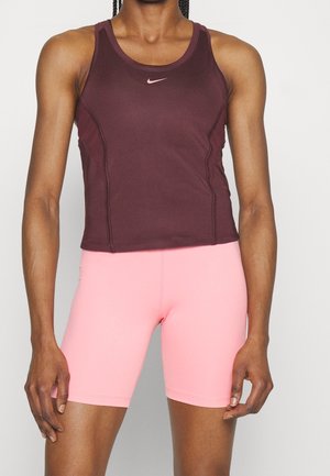 Athlète portant un haut Nike sans manches bordeaux et un cuissard de vélo ajusté rose, debout devant un fond clair uni.