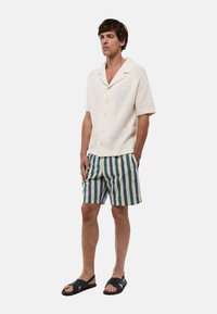 Chemise en tricot blanche à manches courtes avec des trous verticaux, fermeture à boutons ; associée à un short rayé vert, bleu et beige ; sandales noires.
