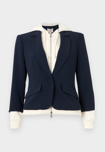 Navy tailored blazer layered over a cream zip-up hoodie with drawstrings, showing hoodie cuffs and hem extending beyond blazer edges.
