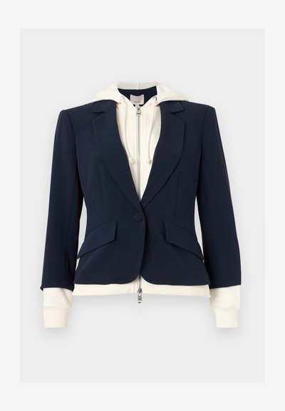 Navy tailored blazer layered over a cream zip-up hoodie with drawstrings, showing hoodie cuffs and hem extending beyond blazer edges.