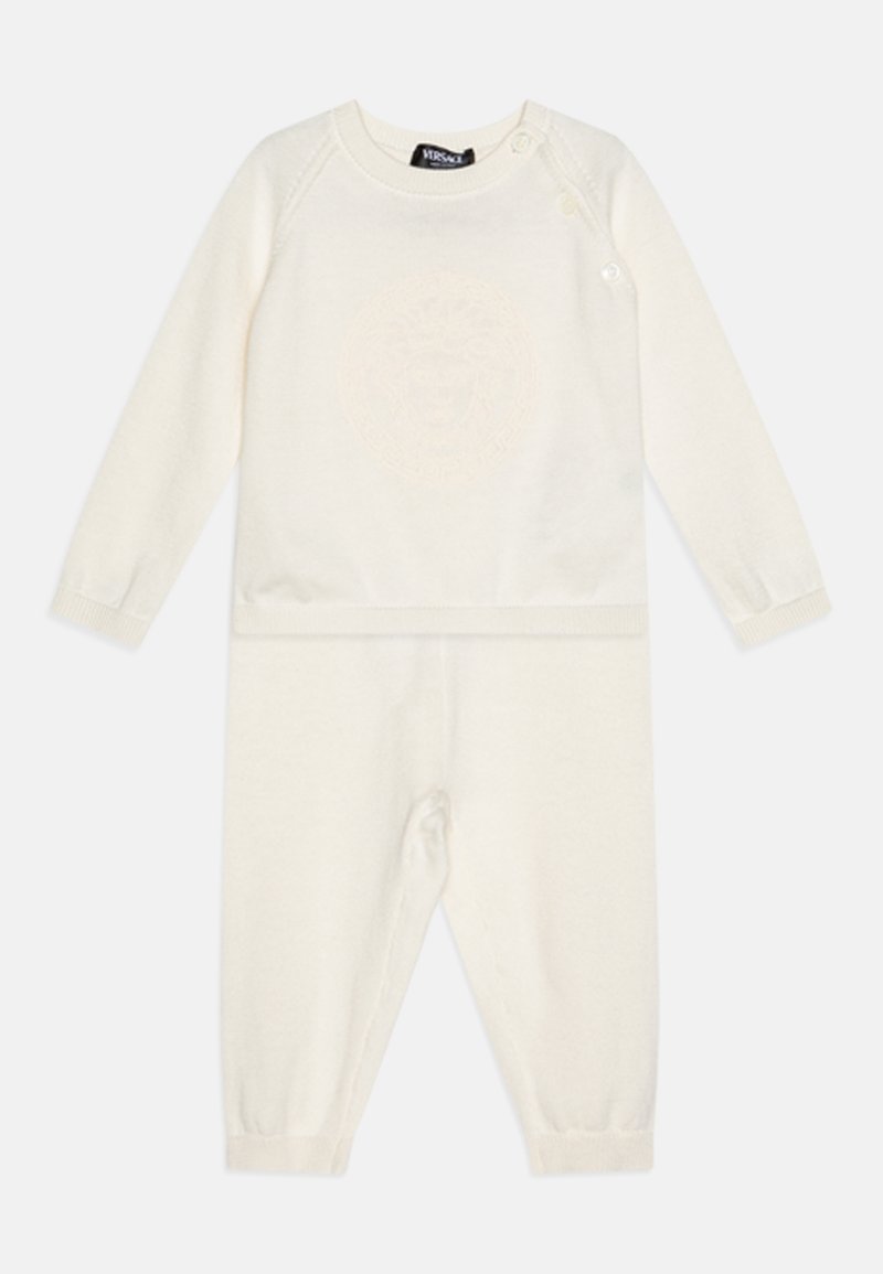 Jumpsuit de punto color crema con mangas largas, que presenta un sutil relieve de un emblema de león, botones a presión en los hombros y puños acanalados.
