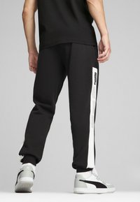 Schwarze Jogginghose mit einem weißen Streifen an der Seite, der das "Puma"-Logo zeigt. Aus weichem Stoff, lässige Passform mit elastischen Bündchen. Getragen mit grauen Sneakers.