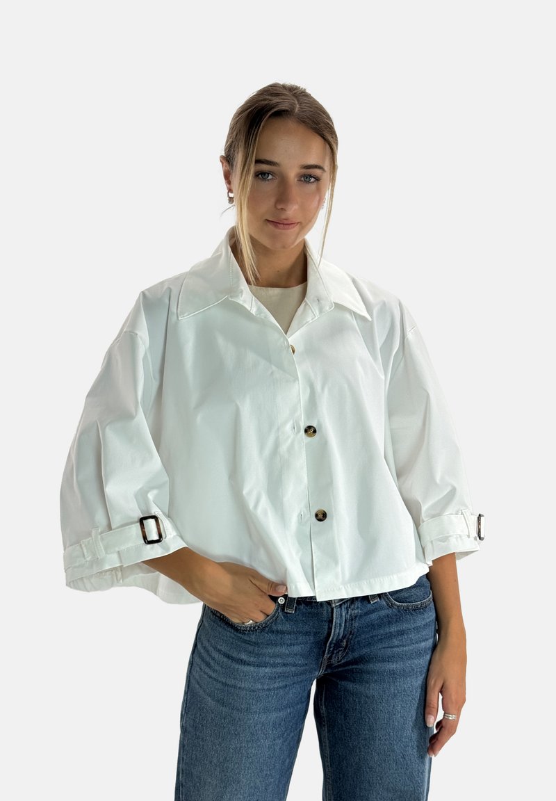 Chaqueta blanca recortada con cuello de solapa, botones delanteros y mangas enrolladas con correas ajustables. Combinada con jeans azules.