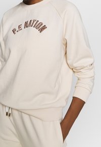 Krämfärgad sweatshirt med raglanärmar, som har "P.E. NATION" broderat i bruna bågformer, ribbade ärmslut och en mjuk textur.