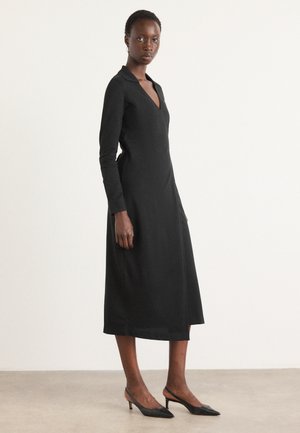 Black, long-sleeve dress with a V-neck and collar, structured fit, knee-length, made of smooth fabric. Paired with black pointed heels.