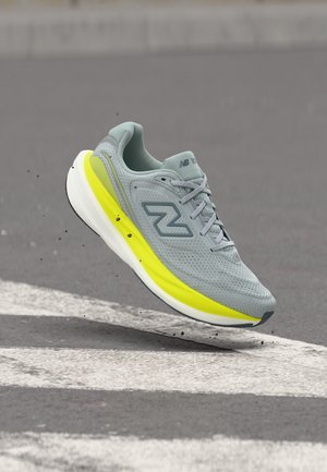 Light gray running shoe with yellow sole midsection, suspended above asphalt with white road markings and small black debris.