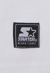 Svart rektangulær etikett på hvit stoff, med "STARTER" i stilisert skrift og en stjernegrafikk, med "BLACK LABEL" under i hvit tekst.