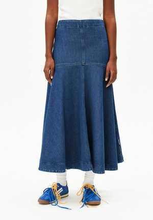 Personne portant une longue jupe en jean, un haut blanc, des chaussettes blanches et des baskets bleu vif à lacets jaunes, debout devant un fond uni.