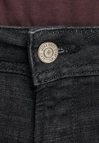 Jeans de mezclilla negra con un acento de botón plateado y logo en relieve, que presentan un acabado texturizado y detalles cosidos a lo largo de la cinturilla.
