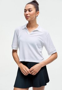 Bela skrajšana polo majica z ravnim ovratnikom in kratkimi rokavi, kombinirana s črno naborkano krilo. Gladka tekstura, minimalističen dizajn.