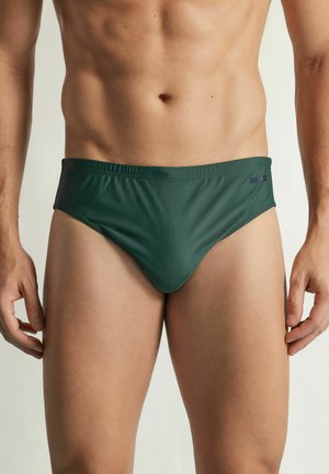 Mannelijke torso met donkergroene zwembroek, staand met ontspannen armen langs het lichaam tegen een effen achtergrond.