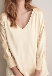 Blouse couleur crème avec un décolleté en V et des manches trois-quarts. Fabriquée en tissu lisse et brillant avec une coupe décontractée et des poignets ajustés.