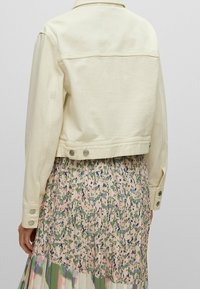 Krátká krémová džínová bunda s dlouhými rukávy, zdobená kovovými knoflíky a s kratším střihem, předvedená přes maxi šaty s květinovým vzorem.