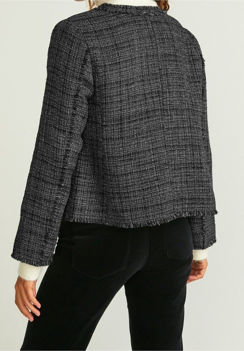 Schwarze strukturierte Jacke mit ausgefransten Kanten und einem kastigen Schnitt. Lange Ärmel und ein einfaches Kragen-Design. Über dunklen Hosen getragen.