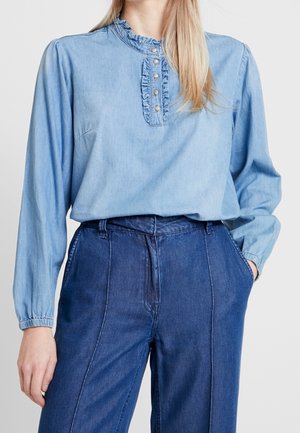 Helle blaue Denim-Bluse mit Rüschensaum und Knopfleiste mit sieben Knöpfen. Lange Ärmel mit elastischen Bündchen. Kombiniert mit dunkelblauen, hüfthohen Hosen.