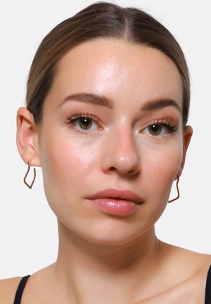 Orecchini geometrici color rame presentano un design ondulato e aperto. Il modello ha un'espressione neutra, con pelle liscia e trucco leggero.