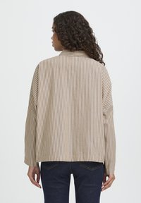 Kvinde med langt krøllet hår står med ryggen til, iført en beige og hvid vertikalt stribet skjorte og mørkeblå jeans.