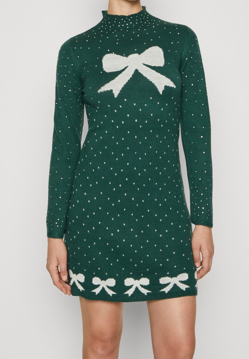 Virkat klänning i mörkgrön med långa ärmar, prydd med ett vitt rosett-mönster på bröstet och vita rosetter längs nederkanten, dekorerad med små silverprickar.
